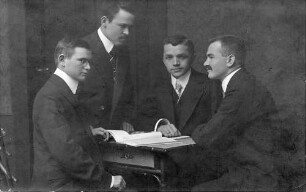 Vier Männer, Schneidergesellen, vorne links Jakob Müller, in Anzügen um einen Schreibpult mit zwei Heften darauf Postkarte mit Briefmarke vom 30.05.1912 an Babette Müller, Ellerstadt, von Jakob Müller aus Paris