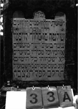 Grabstein 33 (Index: 100101442) Material: Sandstein, weiße Marmorplatte, Abschuß Kupfer Zustand: Sockel mehrfach gesprungen und verwittert Maße HxBxT: 140x42x38 cm Beschreibung: rechteckige Stele, Sockel, achteckiger abgeflachter Abschluß, Schriftplatte mit dreibogigem Abschluß: Grabstein 33A