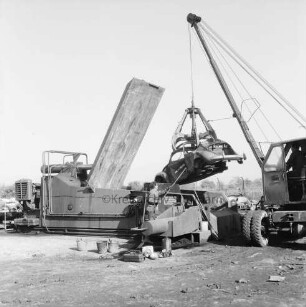 Harry Kloss Autoverwertung GmbH & Co. KG: Kampstraße: Abstellplatz mit Schrottautos: rechts Bagger beim Hochheben eines VW-Käfers mit Greifer: hinten Mitte Schrottpresse: daran Arbeiter mit Stange