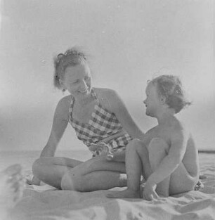 Frau in Bademode und nacktes Kind sitzen am Strand