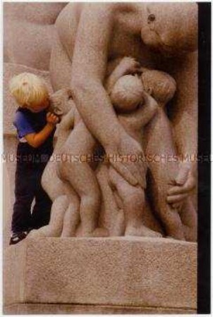 Kleines Kind lehnt neben einer Skulpturengruppe, die eine Gruppe von Kindern und eine, sie alle umarmende weibliche Gestalt zeigt (Altersgruppe 14-17)