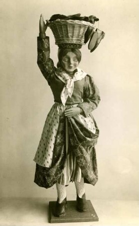 Spielfigur aus Holz in Stoffkleidung. Frau mit auf dem Kopf gestützen Korb. Schuhändlerin