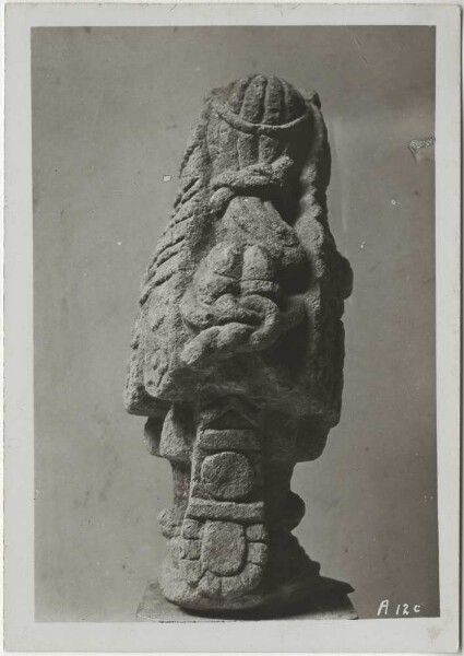 "View of a very peculiar stone figure (light grey Tezontli). "... Head wearing rich floral jewellery, right hand bouquet, left hand flower (calla). Ear jewellery of Macuilxochitl, bell belt, pubic bandage, sandals, face and torso (skeleton!) of the god Xipe"