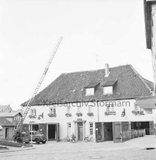 Hude: Feuerwehrgerätehaus "Blaues Haus" mit geöffneten Toren: links Feuerwehrfahrzeug mit ausgefahrener Drehleiter: in der Mitte Mädchen mit Kinderwagen und Fahrräder