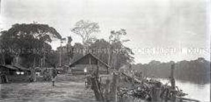 Ansicht eines Dorfes am Flussufer mit einem afrikanischen Angehörigen der Polizeitruppe und einem Kolonialbeamten im Hintergrund