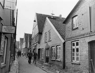 Otterndorf an der Medem. Stadtansicht. Kleine geduckte Backsteinhäuschen rechts und links der Straße auf der sich Passanten befinden. Im Vordergrund eine Autovermietung