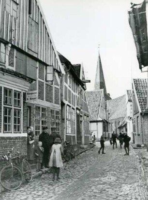 Otterndorf an der Medem. Straßenszene. Kleine geduckte Backsteinhäuschen rechts und links der Straße auf der sich Schulkinder befinden. Im Vordergrund eine Klempnerei