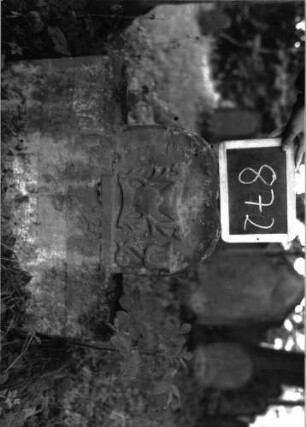 Grabstein 872 (Index: 100102983) Material: Sandstein Zustand: stark verwittert, eingesunken, vollständiger Textverlust Maße HxBxT: 84x76x12 cm Beschreibung: Stele, eingezogener Rundbogenabschluss, im Bogenfeld stilisierte Levitenkanne (Kelch), von Blumen eingerahmt (eine abgewittert) Symbolik: Levitenkanne ?, Blumen: Grabstein 872