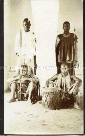 Gruppenbild von vier Kriegern der Kaka und Jangere, mit Waffen und einer Trommel, vor neutralem Hintergrund