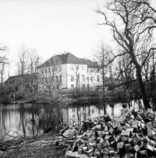 Gut Altfresenburg: Herrenhaus, davor der Gutsteich, vorne rechts Person hinter aufgehäuften Holzstücken