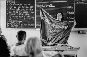 Geschichtsunterricht an einer Realschule. Die Lehrerin hält eine Fahne der DDR Jungen Pioniere hoch. Seid bereit, immer bereit