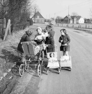Dorfstraße: vorn Mädchengruppe mit Puppenwagen: dahinter Junge: links Hecke und Büsche: hinten Bauernhäuser