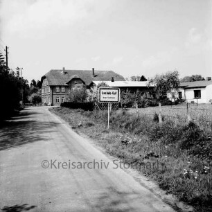 Lasbek-Gut: Ortseingangschild: vorn links Lasbeker Straße: rechts eingezäunte Weide: hinten Bauernhäuser und Bäume