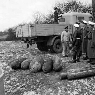 Bombenräumung: Wiese zwischen Turmstraße und Industriestraße: vorn vier entschärfte 500-Kilogramm-Bomben und eine 1000-Kilogramm-Bombe: rechts auf Boden Gestänge des Dreibeins: dahinter der Leiter der Polizeiinspektion Bad Oldesloe, Polizeirat Werner Schedalke, sowie drei Polizeibeamte und ein Arbeiter: hinten Lkw: darauf Bernd Althausen vom Bombenräumkommando Kiel [später: Kampfmittelräumdienst Kiel]: dahinter Büsche: im Hintergrund rechts Teil des Wasserturms: Winter
