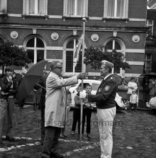 Oldesloer Spielmannszug e.V.: Markt: Entgegennahme der Standarte: Vorsitzender des Bürgervereins Alwin Thies übergibt Standarte an Dirigent Paul Fedder: im Beisein von Stadtverordnetem Georg Wolfgang Dittmer: zwei Jungen schauen zu: hinten parkende Autos, Zuschauer