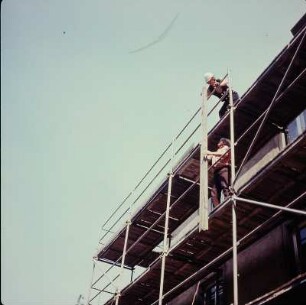 Zimmerer auf einem Gerüst beim vertikalen Weiterreichen einer Holzbohle