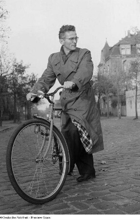 Ein Mann auf einem Fahrrad präsentiert einen Trenchcoat des VEB Bekleidungswerk Leipzig