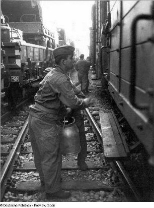 Deutschland . Truppenübungsplatz Klietz. Nationale Volksarmee - Landstreitkräfte. Truppenverlegung per Eisenbahnmarsch. Soldaten zwischen zwei Militärzügen (links Flachwagen mit Militärtechnik, rechts Mannschaftswagen). Im Vordergrund Soldat mit Milchkanne (für Waschwasser?)