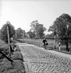 Drahtmühle: Kopfsteinpflaster: rechts Frau auf Fahrrad: dahinter Brücke über Mühlenbach: links und rechts Verkehrsschilder: hinten Weiden und Bäume