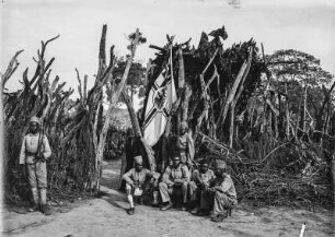 Deutsche Kolonialsoldaten : Deutsche Kolonialsoldaten am Eingang zu einer Boma. An der Umzäunung wurde die Kaiserliche Kriegsflagge gehisst