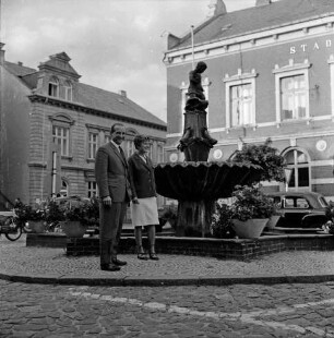 Marktplatz: vorn links Paar vor Gänselieselbrunnen mit Blumenschmuck: dahinter Pkws, Motorräder, Stadthaus und Wohnhaus an Hagenstraße