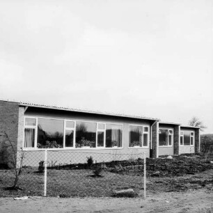 Volksschule: Neubau: Außenansicht: davor unbefestigte Außenanlagen und Zaun