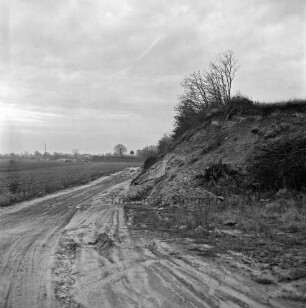 Feldmark: Wirtschaftsweg: rechts Hang von Kiesgrube mit Buschwerk: hinten links Sandhaufen, Lkw und Schornstein hinter Bäumen