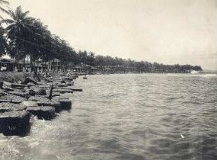 Colón, Panama. Blick in die Bucht entlang des Palmenstrandes mit großen Steinblöcken als Wellenbrecher, im Hintergrund Wohnhäuser oder Strandhäuser