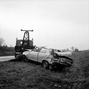 A 1: bei Stapelfeld: Verkehrsunfall: Pkw rammt Pkw und prallt gegen Brückenpfeiler: schräg neben Fahrbahn Unfall-Pkw, auf Straße Abschleppwagen, davor liegender Mann beim Befestigen des Hakens an Autowrack: hinten durchfahrende Autos, Hinweisschild auf Autoabfahrt Stapelfeld, Zäune