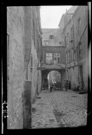 Nicht identifizierbare Ortschaft bei Saint-Quentin (?) (Dep. Aisne, Frankreich): Quergebäude mit Torbogen über einer gepflasterten Ortsstraße, davor Soldaten neben einem Pferdegespann (Distanzaufnahme)
