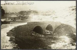 Steinbrücke in einem Tal des Taurusgebirges, im Hintergrund nähert sich ein Fahrzeug auf der Straße, Umrisse eines Zeltlagers am Fuße eines Hügels