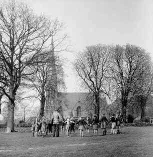 Friedenskirche: Außenansicht von Süden: vorn Dorfanger: darauf Schulkinder mit Lehrer beim Spiel: dahinter Kirchhof mit Bäumen