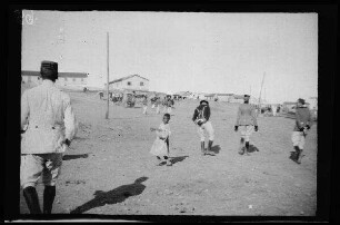 "Maurenstadt" neben dem Lager von Nador (Marokko): Einheimische Kinder und Offiziere [des spanischen 8. Kavallerie-Regiments "Lusitania"] in hellgestreiften Uniformhosen, dunklen Uniformjacken und niedrigen, runden Feldmützen auf einem Platz mit Gebäuden im Hintergrund [Eröffnung der Bahnstrecke Nador-Zeluan (Selouane, Marokko] (Rückansicht)