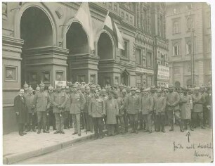 Ca. 60 Personen (grösstenteils Offiziere) teils in Uniform mit Mütze oder Pickelhaube, teils in Zivil vor beflaggtem Gebäude auf Strasse in der Innenstadt Warschaus stehend