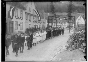 Primizfeier Heinzelmann in Stetten unter Holstein; Prozession durch einen geschmückten Torbogen; im Mittelpunkt weißgekleidete Mädchen; im Hintergrund Musikkapelle