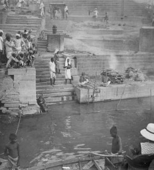 Varanasi (Benares), Indien. Touristen an Deck eines Bootes in Korbmöbeln sitzend, beobachten das Geschehen einer öffentlichen Leichenverbrennung an einem Ghat am Ganges