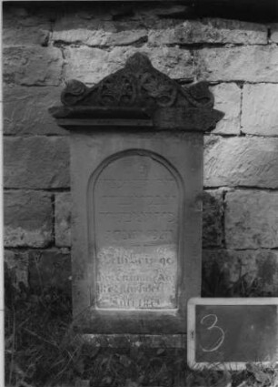Grabstein 3 (Index: 99801813) Material: Sandstein Zustand: verwittert mit Textverlust Maße HxBxT: 82x44x12 cm Beschreibung: Stele, Sockel, Giebel mit Akroteren, eingetieftes Schriftfeld mit Rundbogen Symbolik: Blätterornamentik Besonderheit: Kindergrab: Grabstein 3
