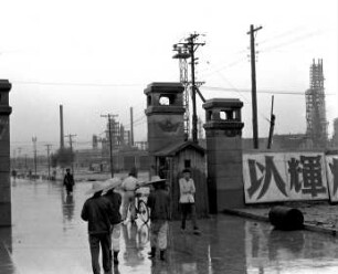 Passanten auf regennasser Straße und Erdölraffinerie im Hintergrund