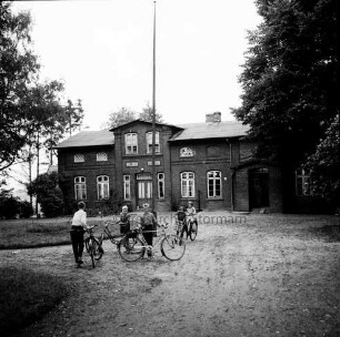 Volksschule: vorn links auf Auffahrt eine Gruppe Mädchen und Jungen mit Fahrrädern: rechts und links Bäume