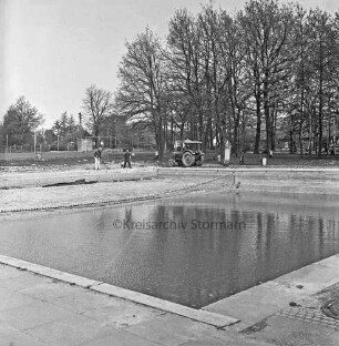 Freibad: vorn Schwimmbecken: dahinter Arbeiter mit Schaufel und Trecker bei Außenarbeiten: hinten links Sportplatz: hinten rechts Bäume und Häuser