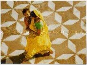 Junge Frau in gelben Sari mit Kind auf dem Arm auf einem geometrisch gepflasterten Platz, von schräg oben gesehen