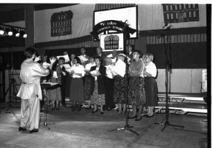 750. Gemeindejubiläum: Abschlussfeier: Sporthalle: Bühne: gemischter Chor beim Singen, mit Dirigentin: Notenständer, Mikrofone, hinten Gemeindeflagge
