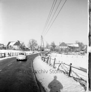 Dorfstraße: links VW-Käfer: rechts Zaun und Weide: hinten Mitte Strommast: im Hintergrund Bauernhäuser und Bäume: Winter