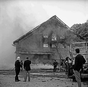Gut Altfresenburg: Brand des Bullenstalls: Brandruine: Giebelfront mit Lastenaufzug: davor Feuerwehrleute, zwei Männer und Wasserschläuche: rechts Teil eines Mähdreschers: hinten Rauchschwaden: 24. August 1971