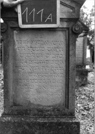 Grabstein 111 (Index: 100080512) Material: Sandstein Zustand: verwittert mit Textverlust Maße HxBxT: 143x47x24 cm Beschreibung: getreppter Sockel, Giebel mit Verdachung, rechteckiges Schriftfeld eingetieft Symbolik: Voluten unterhalb des Giebelansatzes: Grabstein 111A