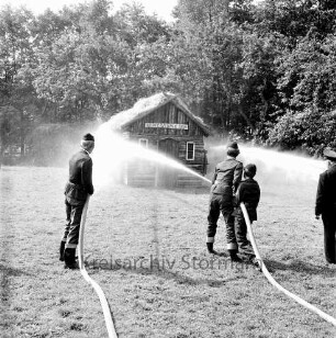 Kupfermühle: Amtsfeuerwehrfest: Löschen der "Kupfermühle Anno 1500": vorn drei Feuerwehrmännder mit Wasserschläuchen: rechts Wehrführer: hinten brennendes Holzhäuschen: dahinter Bäume