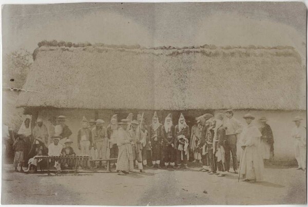 Masked dancers in front of a hut