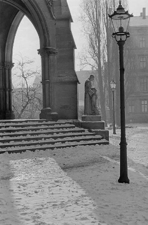 Blick auf einen winterlichen Kirchhof [vermutlich Peterskirche] und die umgebende Bebauung, im Zentrum eine sakrale Sandsteinplastik