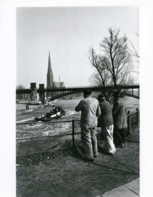 Hamburg-Uhlenhorst. Winter. Eisbrecher fährt in den Kuhmühlenteich vor der Station Uhlandstraße, im Hintergrund St. Gertrud-Kirche. Er bricht eine Fahrrinne. Zuschauer schauen neugierig zu