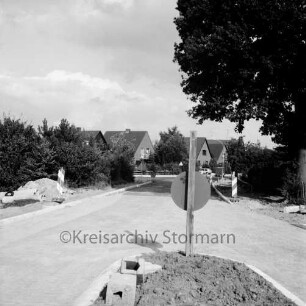 Straßenbau: Oldesloer Straße: Ausbau: Ortseinfahrt: vorn Verkehrsinsel mit Verkehrsschild: rechts und links unbefestigte Fußgängerwege: im Hintergrund Wohnhäuser zwischen Bäumen